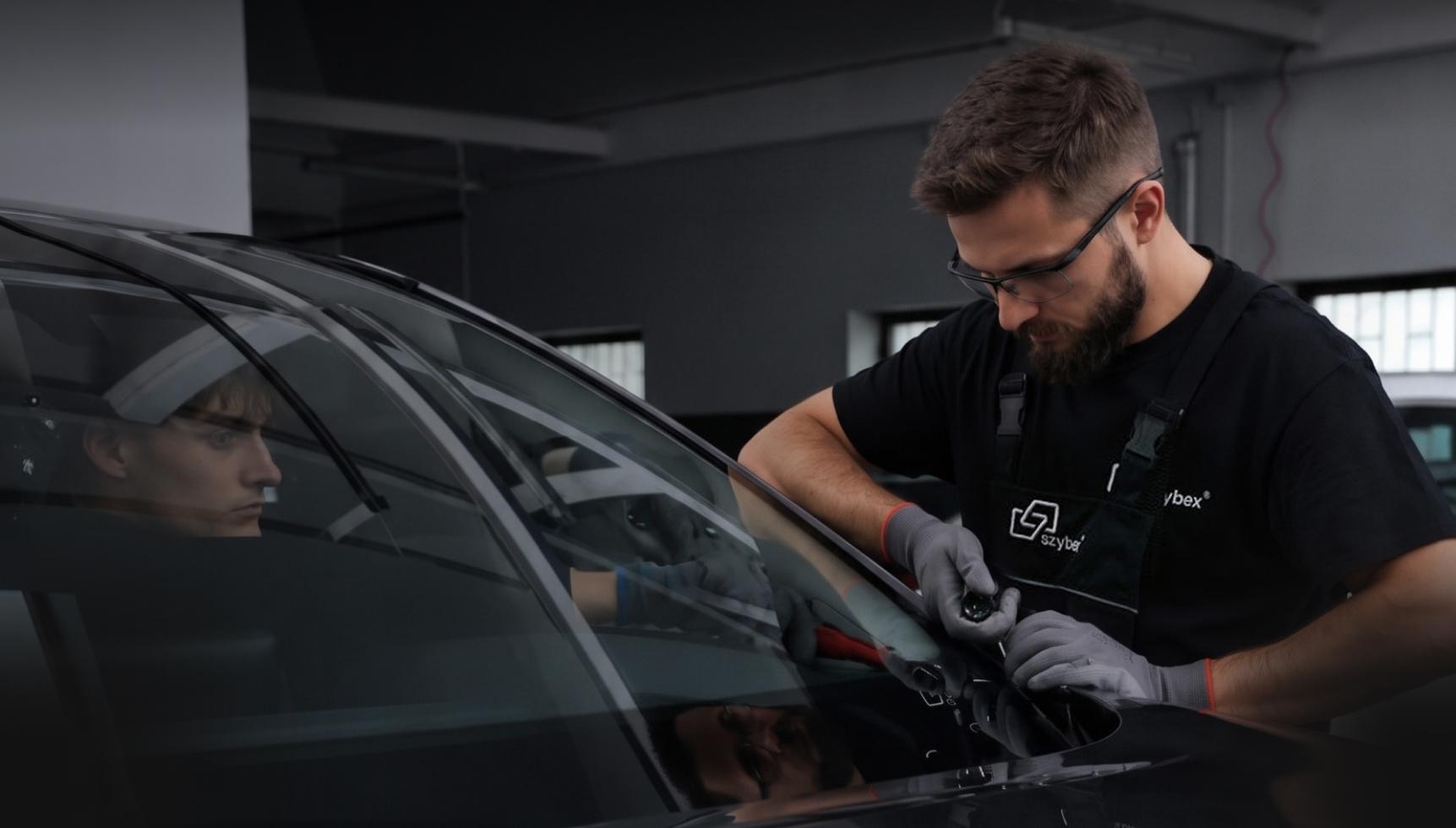 Mechanik w okularach i rękawicach pracuje przy przedniej szybie samochodu, podczas gdy w środku siedzi druga osoba.