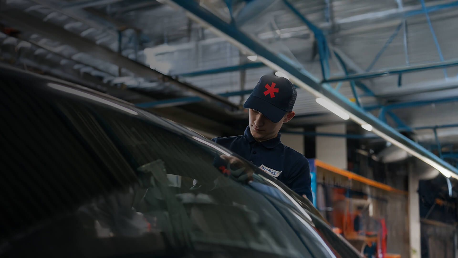 Eine Person mit einer dunkelblauen Kappe mit rotem Symbol arbeitet in einer Werkstatt an einem Auto. Die Umgebung ist industriell geprägt, mit sichtbaren Metallträgern und Geräten.