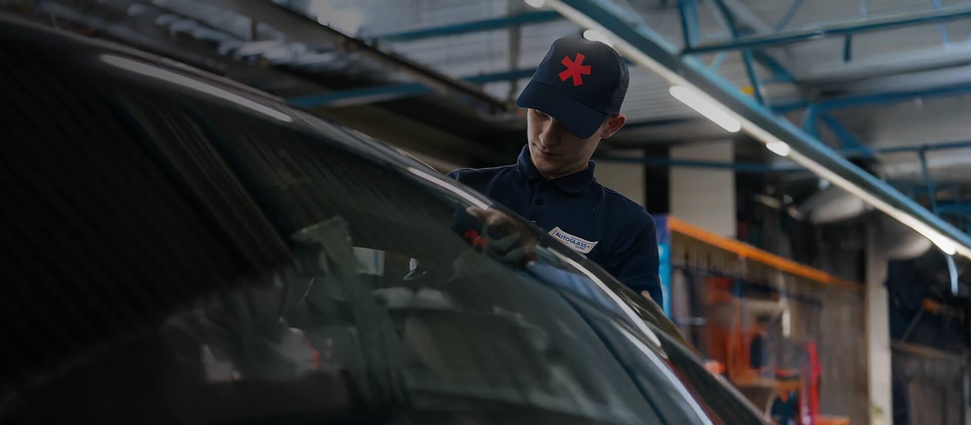 Eine Person mit einer dunkelblauen Kappe mit rotem Symbol arbeitet in einer Werkstatt an einem Auto. Die Umgebung ist industriell geprägt, mit sichtbaren Metallträgern und Geräten.