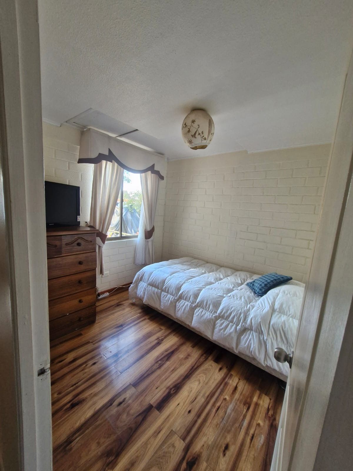 Dormitorio con cama individual, cómoda con televisor antiguo, cortinas y piso de madera.