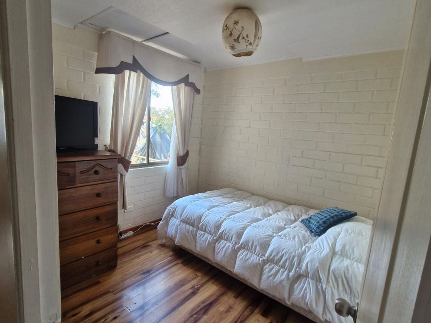 Dormitorio con cama individual, cómoda con televisor antiguo, cortinas y piso de madera.