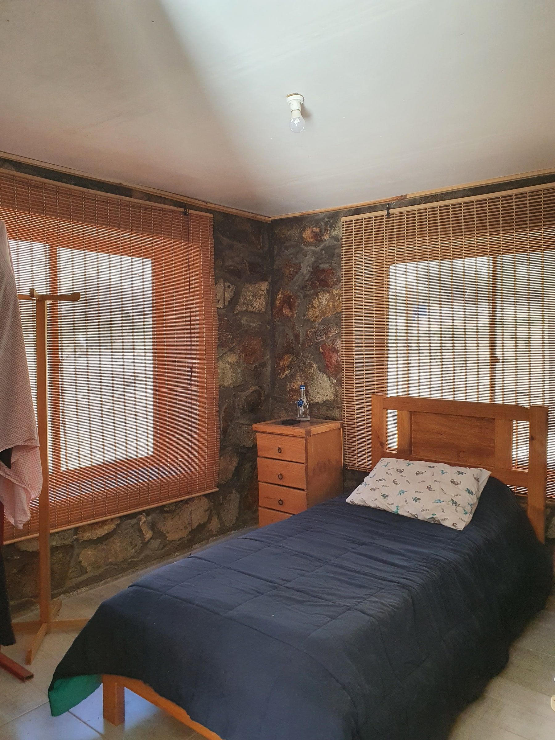Dormitorio con cama, velador y dos ventanas con persianas