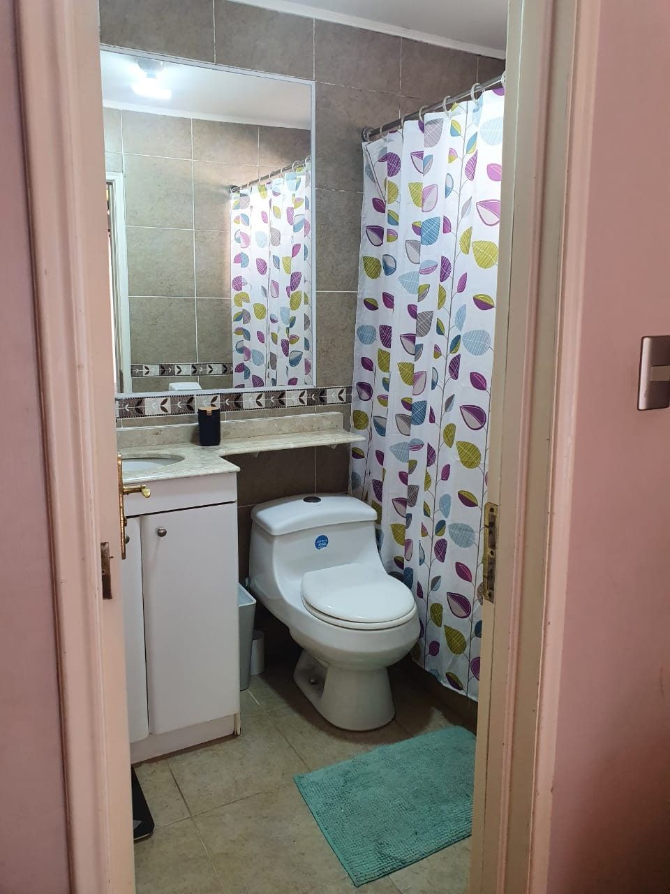 Baño con inodoro, cortina de ducha estampada con hojas y alfombra turquesa en el piso.