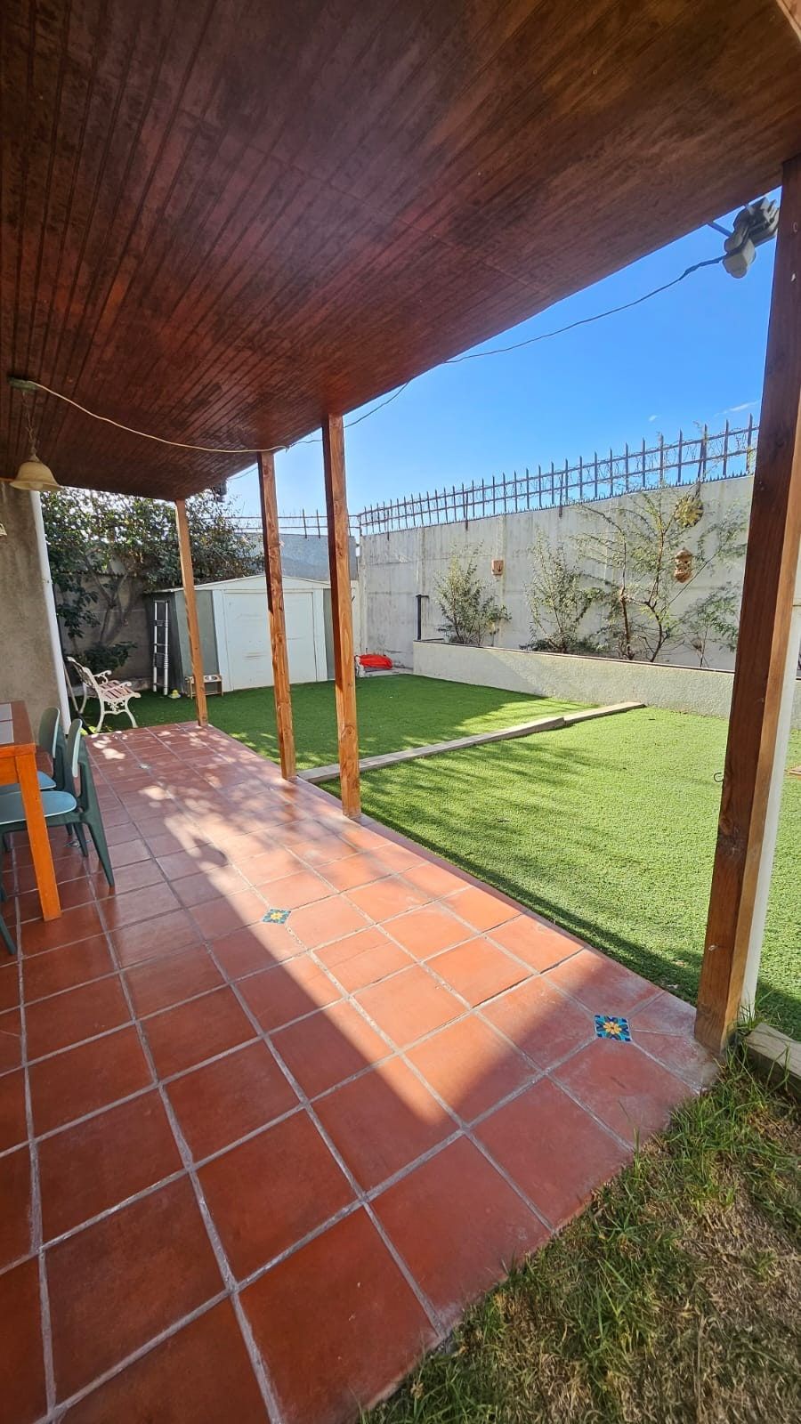 Terraza techada con piso de baldosas rojas, vista al patio con pasto sintético y bodega.