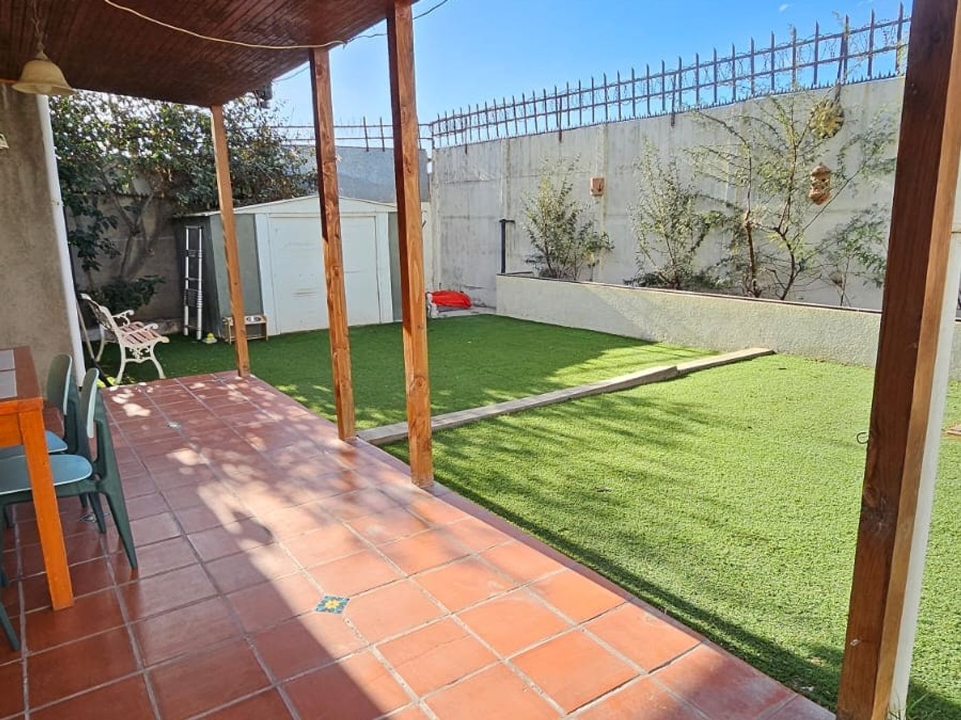 Terraza techada con piso de baldosas rojas, vista al patio con pasto sintético y bodega.
