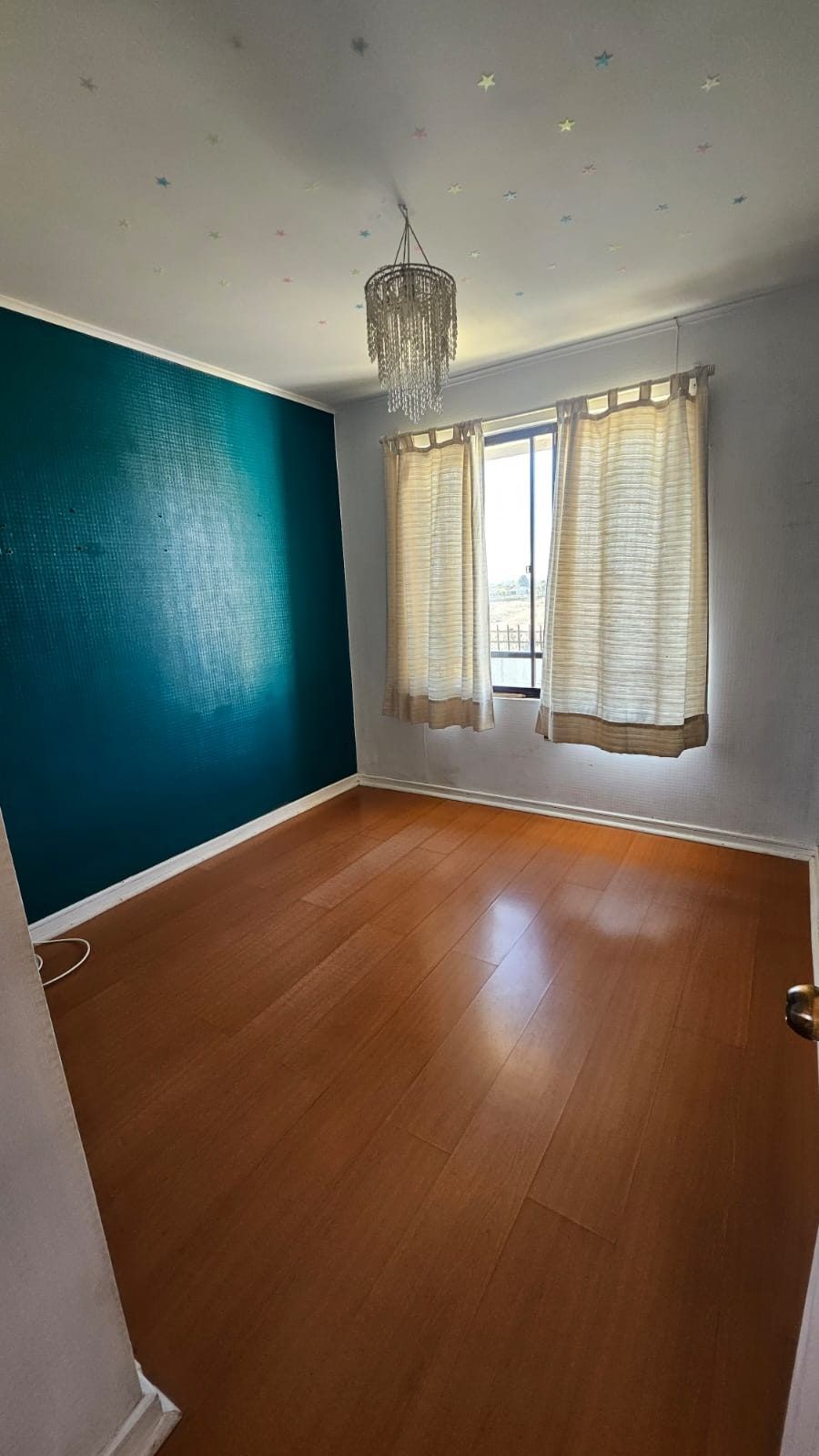 Dormitorio vacío con pared turquesa, piso de madera y lámpara colgante de cristales.