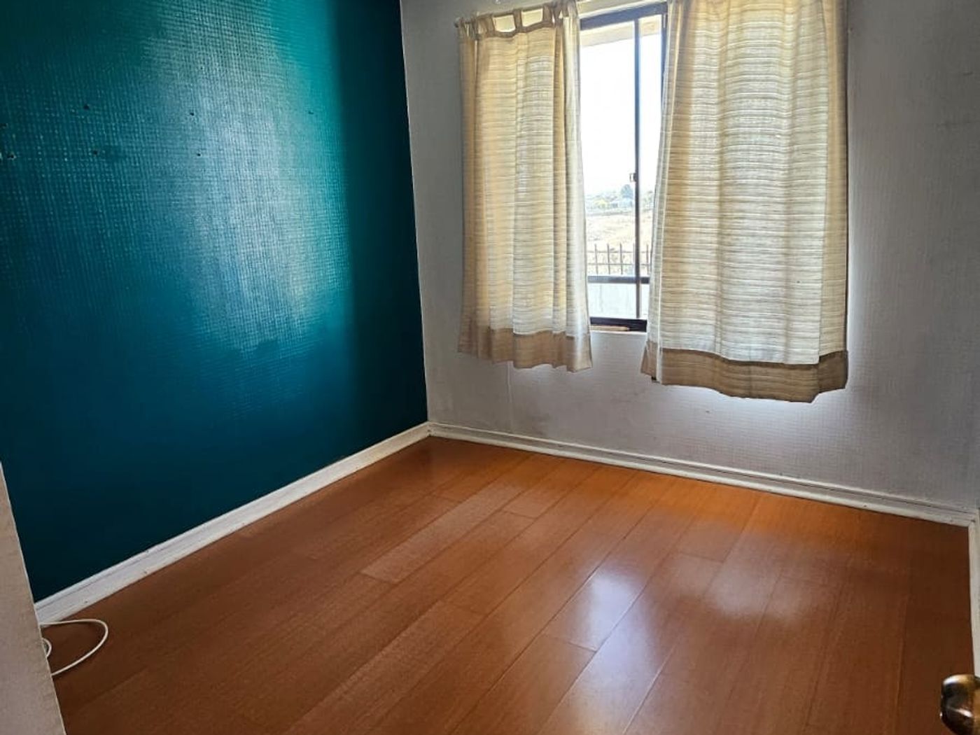 Dormitorio vacío con pared turquesa, piso de madera y lámpara colgante de cristales.