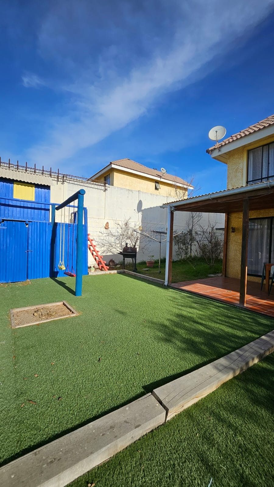 Patio con pasto sintético, juegos infantiles azules y terraza techada al fondo junto a la casa.
