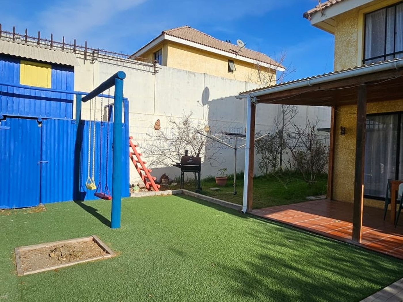 Patio con pasto sintético, juegos infantiles azules y terraza techada al fondo junto a la casa.
