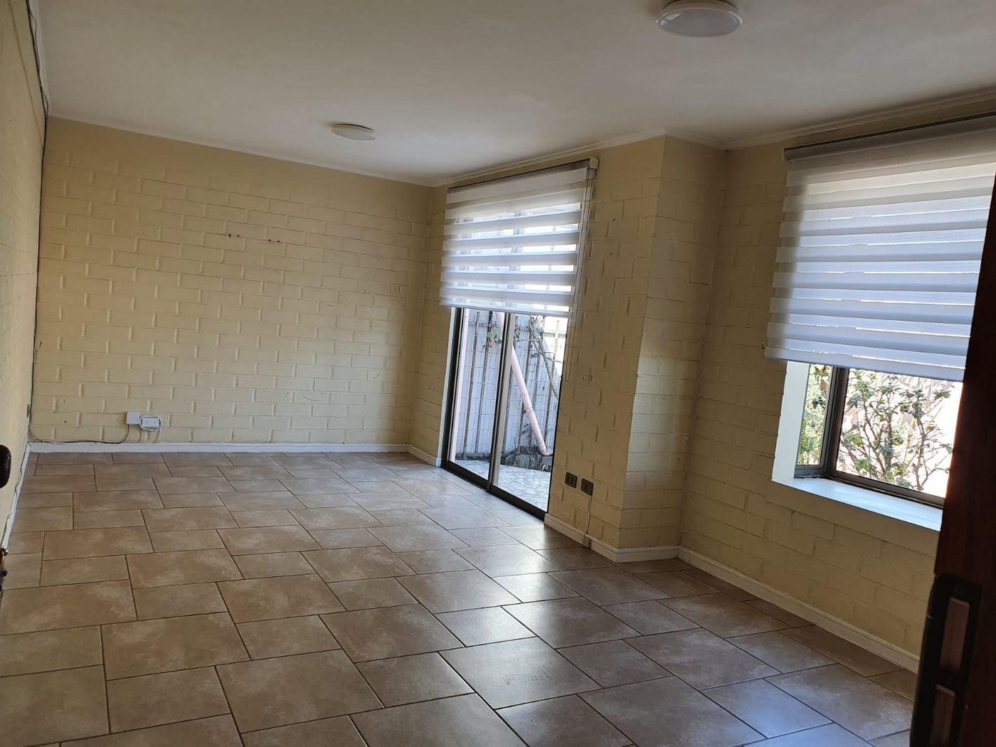 Espacioso living comedor con muros de ladrillo blanco, piso cerámico y dos ventanales con persianas.