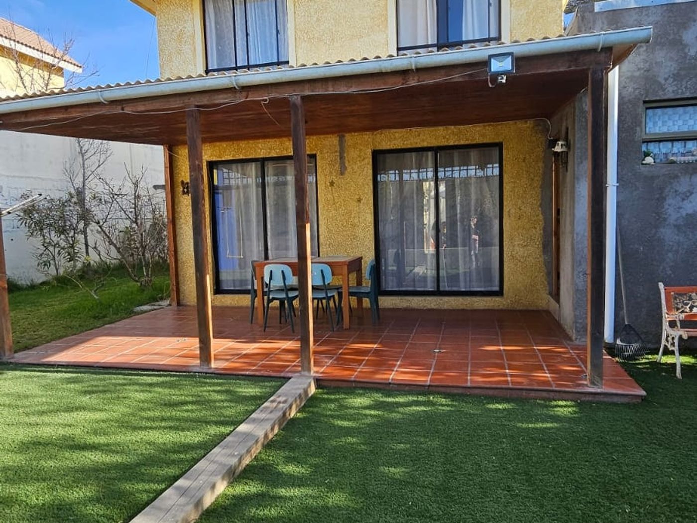 Fachada frontal de la casa amarilla de dos pisos con terraza, comedor exterior y antejardín con pasto sintético.