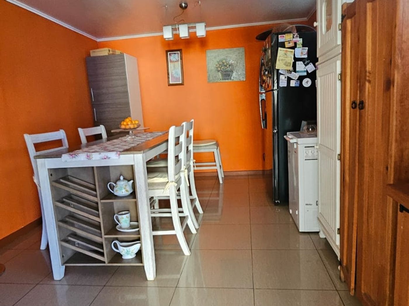 Comedor junto a la cocina con mesa blanca, sillas y refrigerador negro sobre pared naranja.
