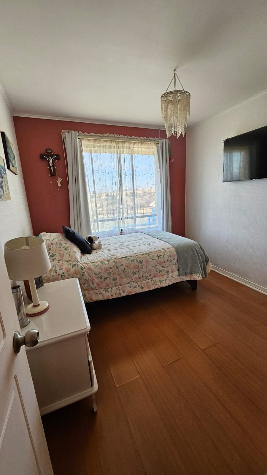 Dormitorio con pared roja, cama, televisor de pared, crucifijo y vista a los cerros.
