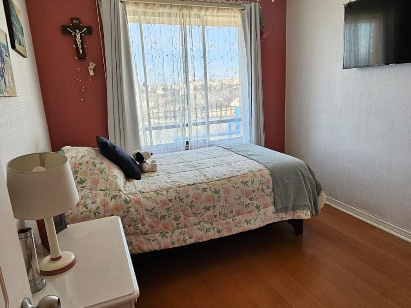 Dormitorio con pared roja, cama, televisor de pared, crucifijo y vista a los cerros.