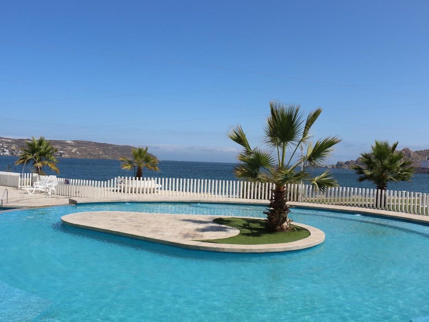 Gran piscina curva del condominio rodeada de palmeras y áreas verdes bajo cielo despejado.