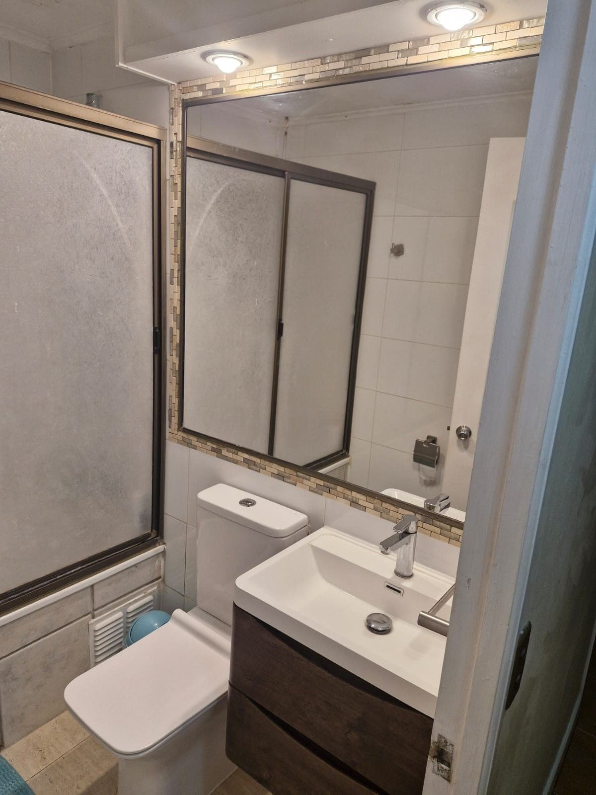 Baño con lavamanos, espejo, estanque de inodoro y mampara de vidrio esmerilado para la ducha.