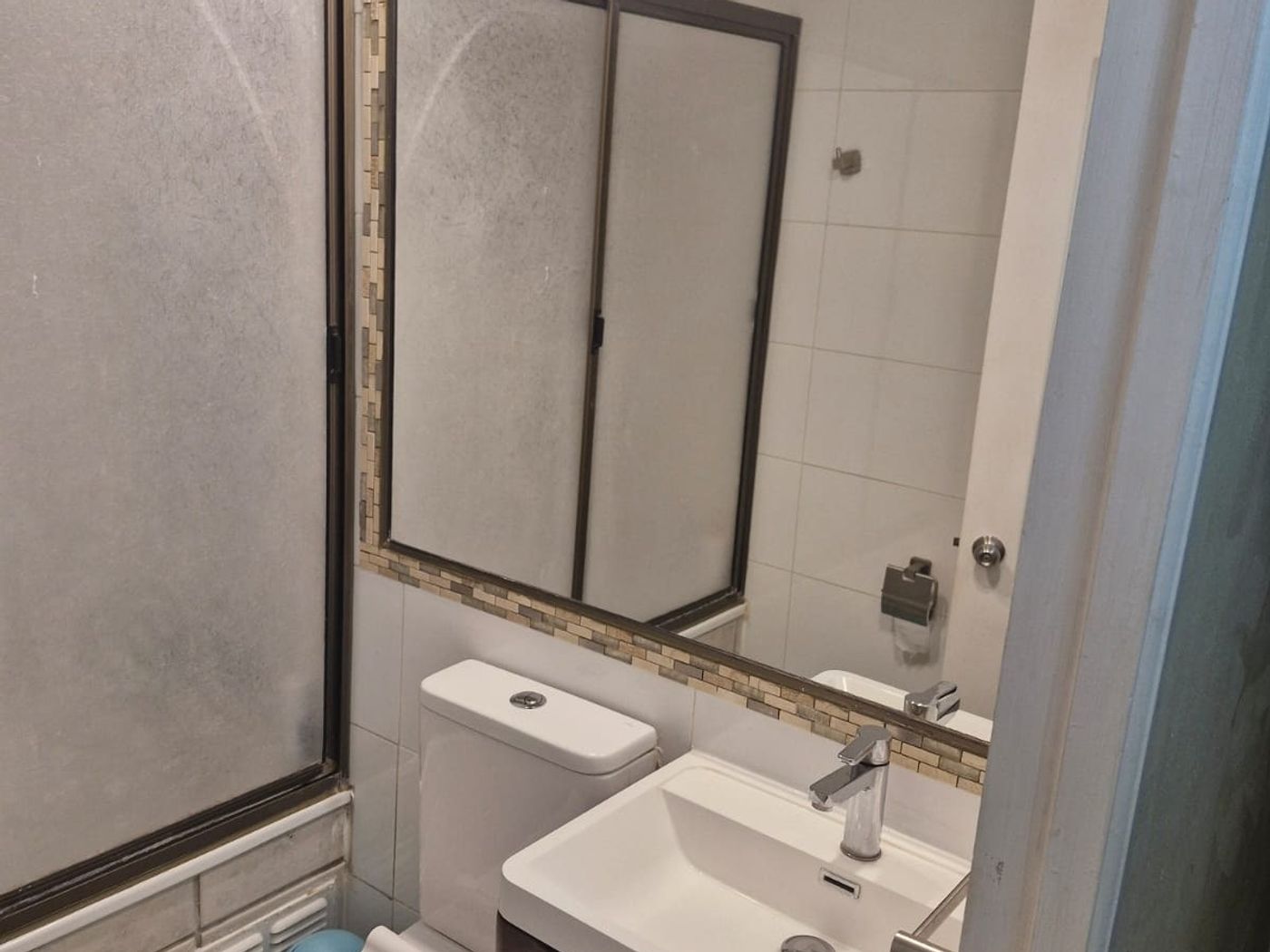 Baño con lavamanos, espejo, estanque de inodoro y mampara de vidrio esmerilado para la ducha.