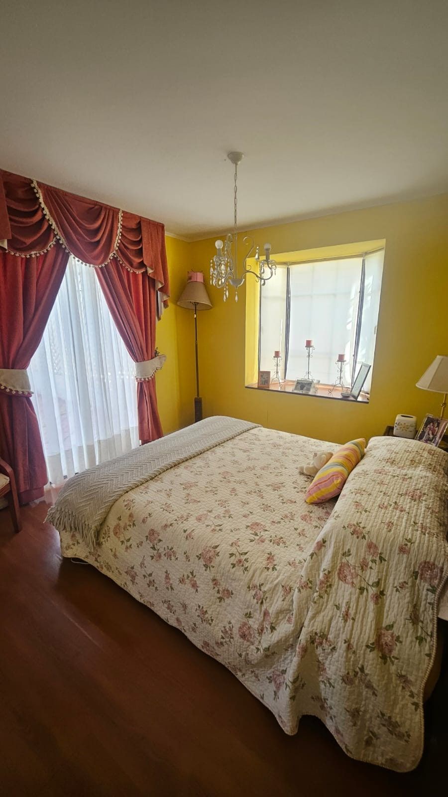 Dormitorio de muros amarillos, cortinas rojas, cama con cubrecama floral y lámpara de araña.
