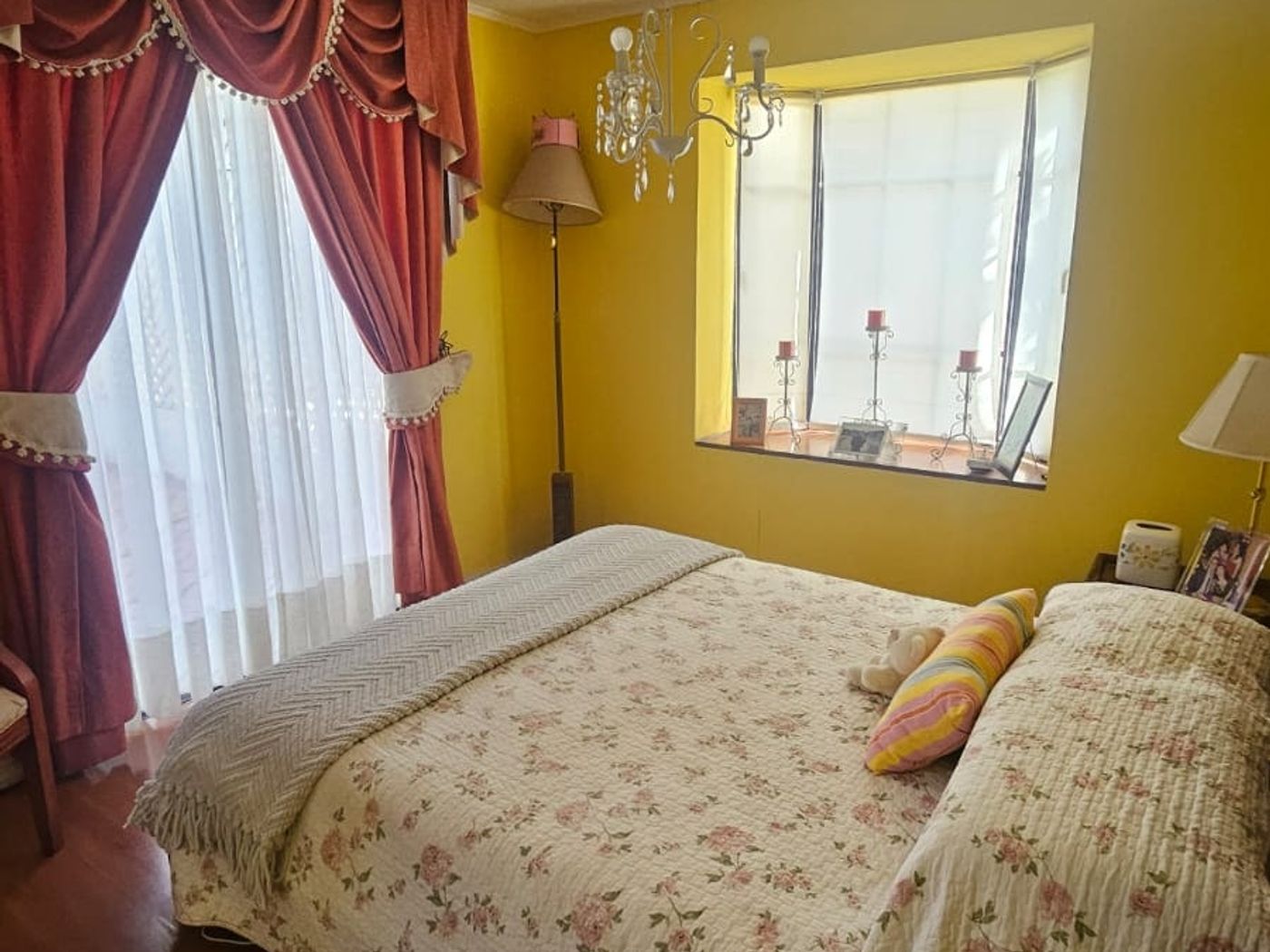 Dormitorio de muros amarillos, cortinas rojas, cama con cubrecama floral y lámpara de araña.