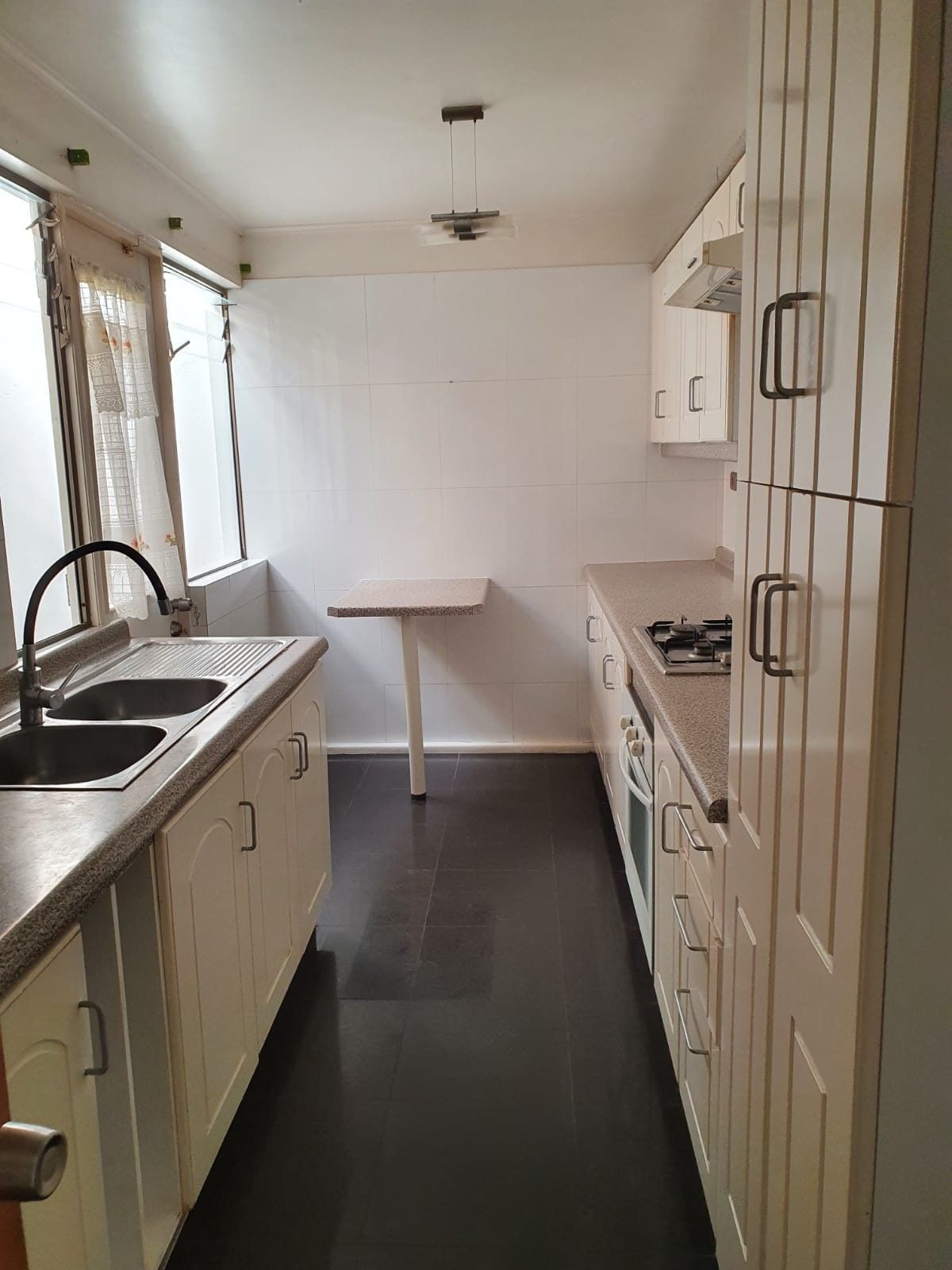 Cocina alargada con muebles blancos, mesón lavaplatos, cocina a gas empotrada y ventana exterior.