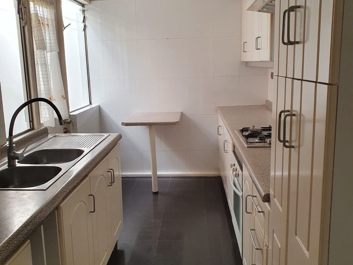 Cocina alargada con muebles blancos, mesón lavaplatos, cocina a gas empotrada y ventana exterior.