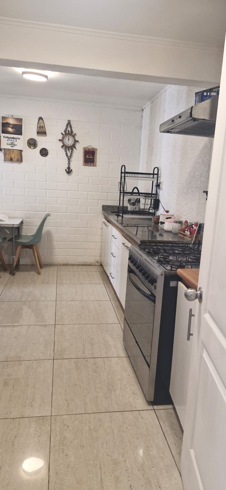 Cocina con pared de ladrillo pintado blanco, cocina a gas, mesa pequeña y reloj decorativo.