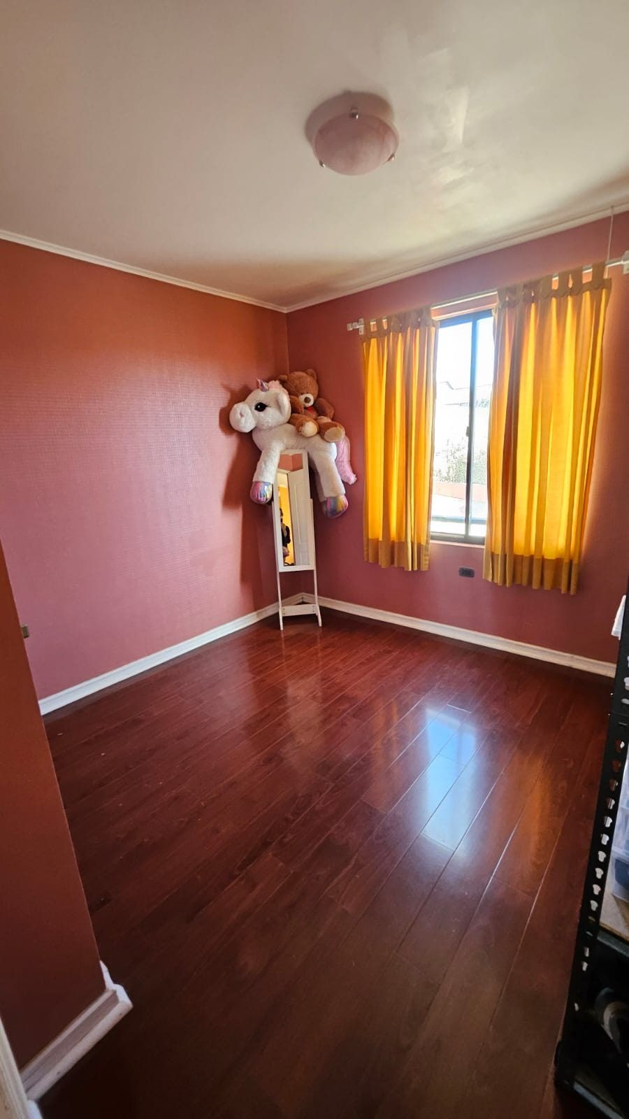 Dormitorio vacío con pared rosada, cortinas amarillas, piso de madera y peluches en un rincón.