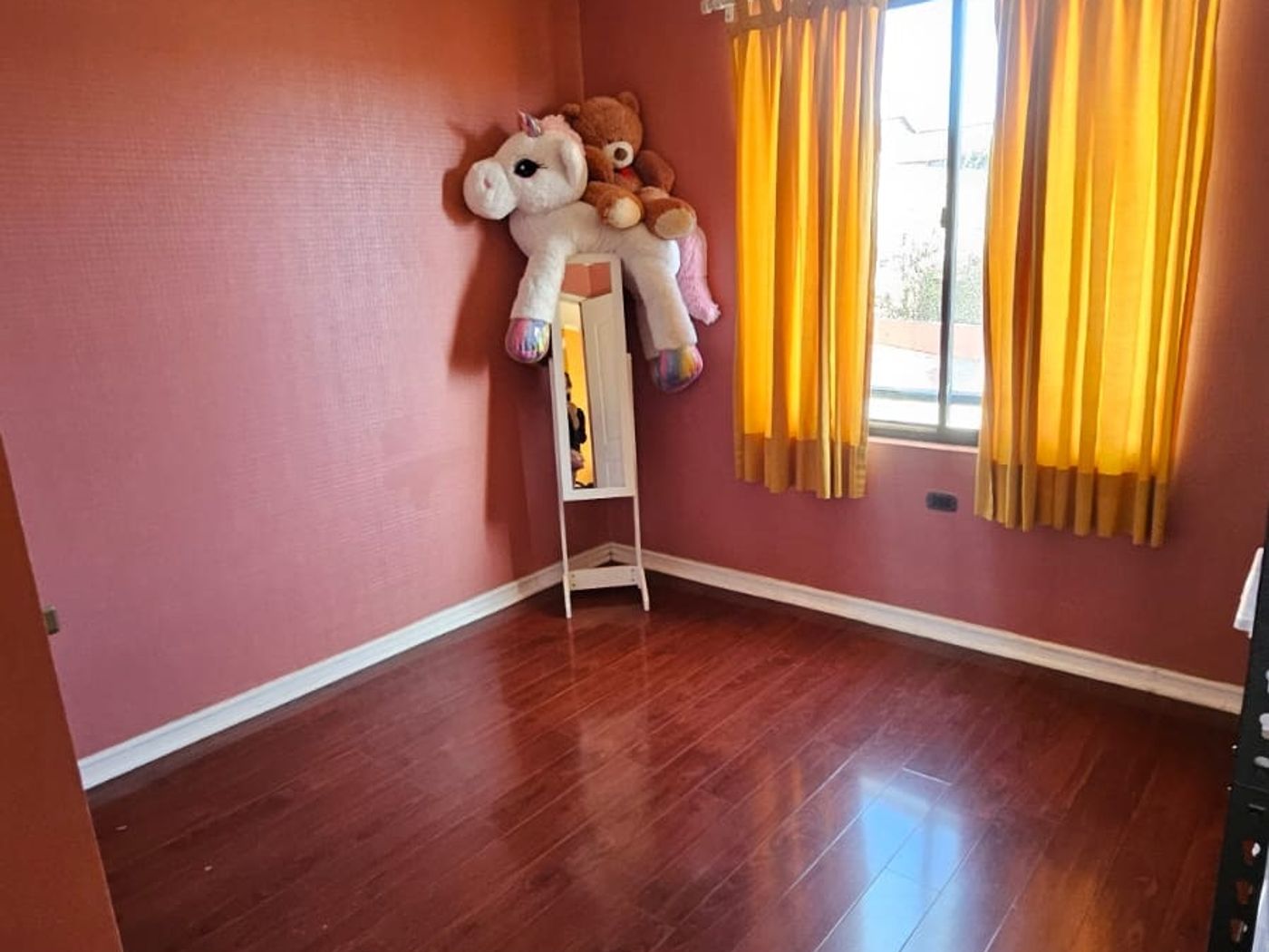 Dormitorio vacío con pared rosada, cortinas amarillas, piso de madera y peluches en un rincón.
