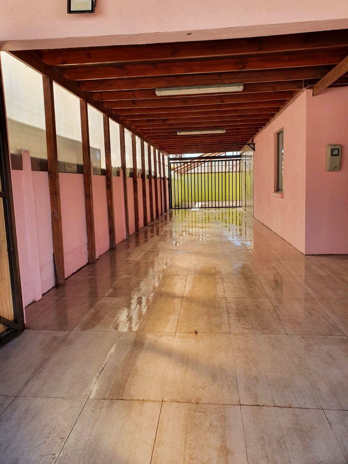 Estacionamiento techado lateral con vigas de madera a la vista y piso cerámico brillante.
