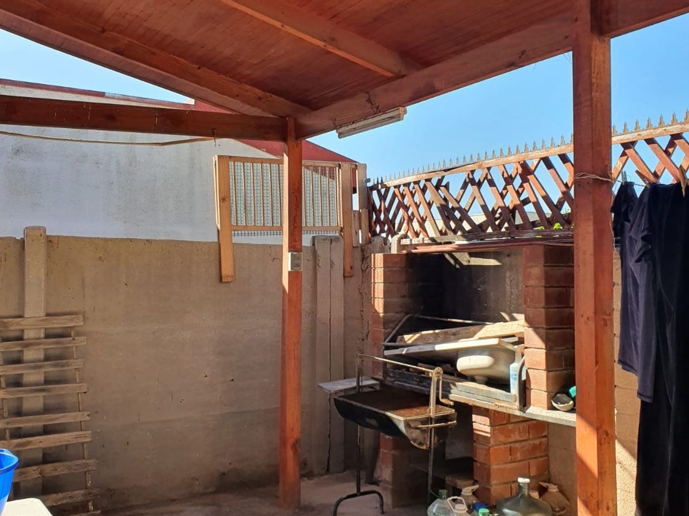 Quincho techado con parrilla de ladrillo, silla, mesa y bidones de agua alrededor.