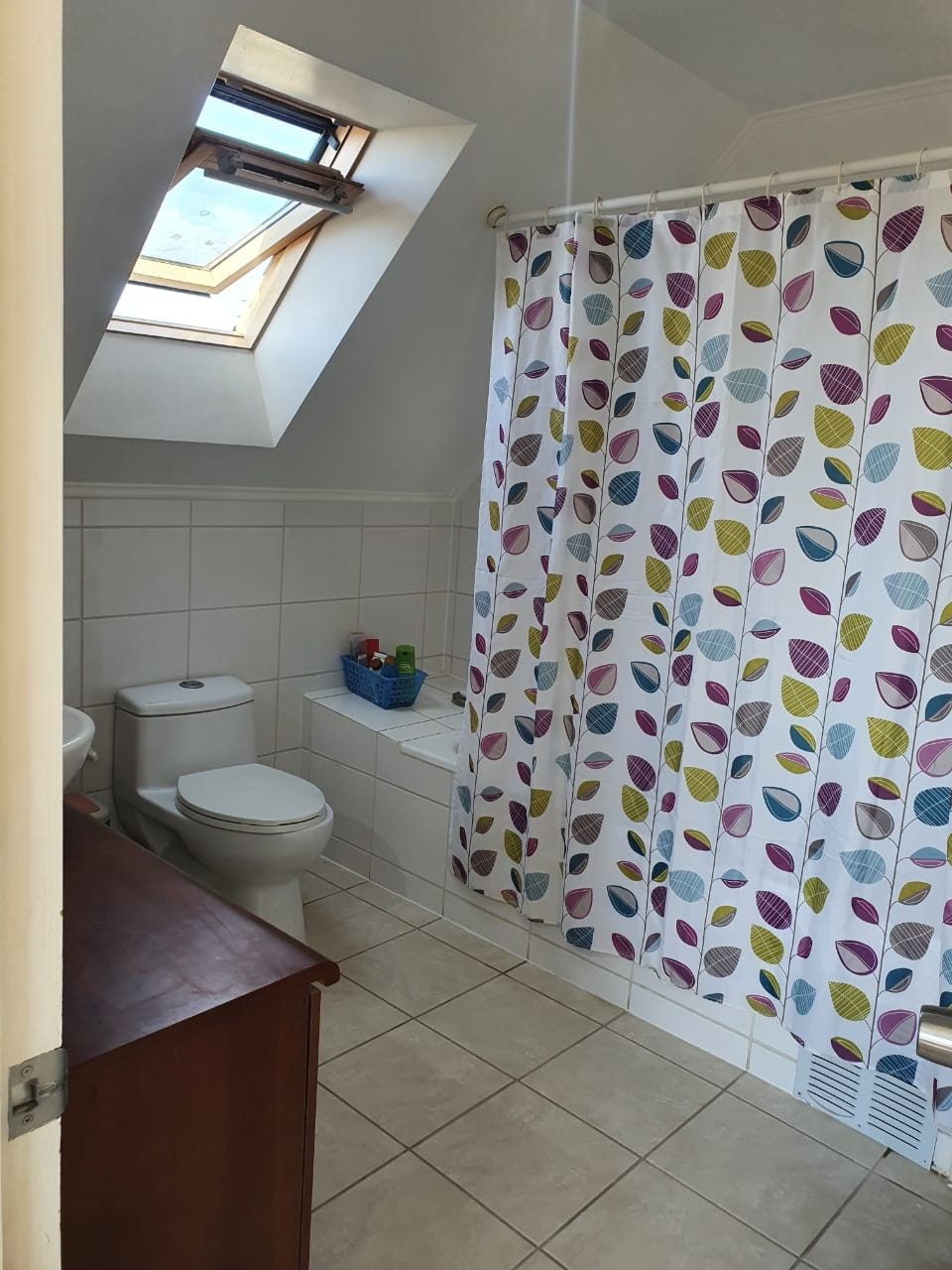Baño con ventana de techo abierta, inodoro, cortina de baño estampada y mueble de madera.