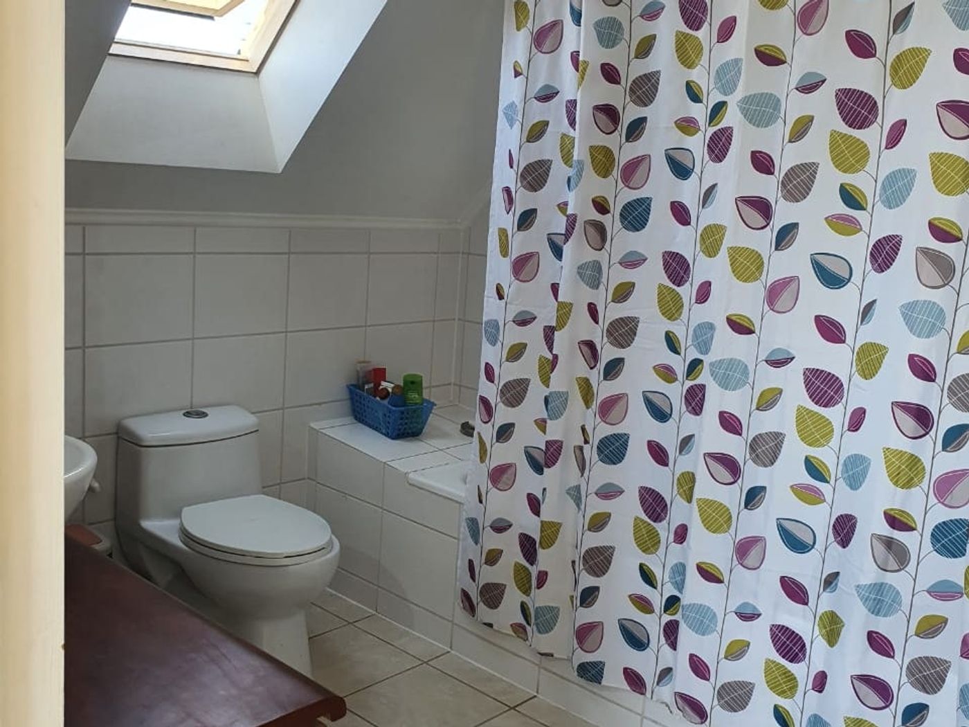Baño con ventana de techo abierta, inodoro, cortina de baño estampada y mueble de madera.