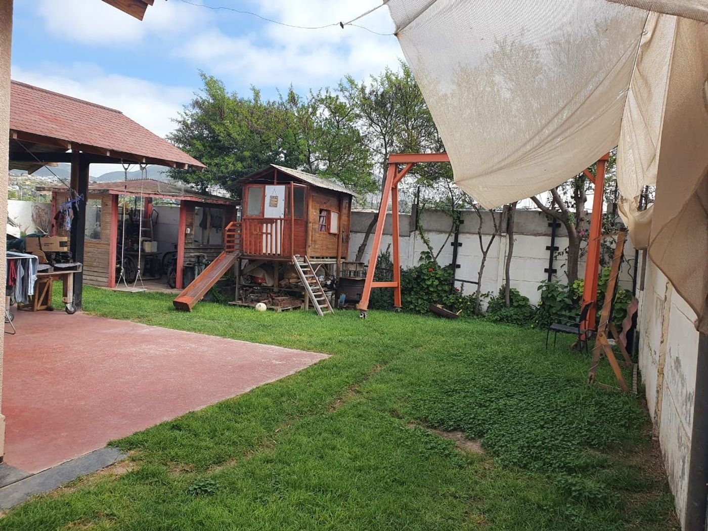 Patio con pasto, quincho techado, casa de muñecos de madera, tobogán y estructura de columpio.