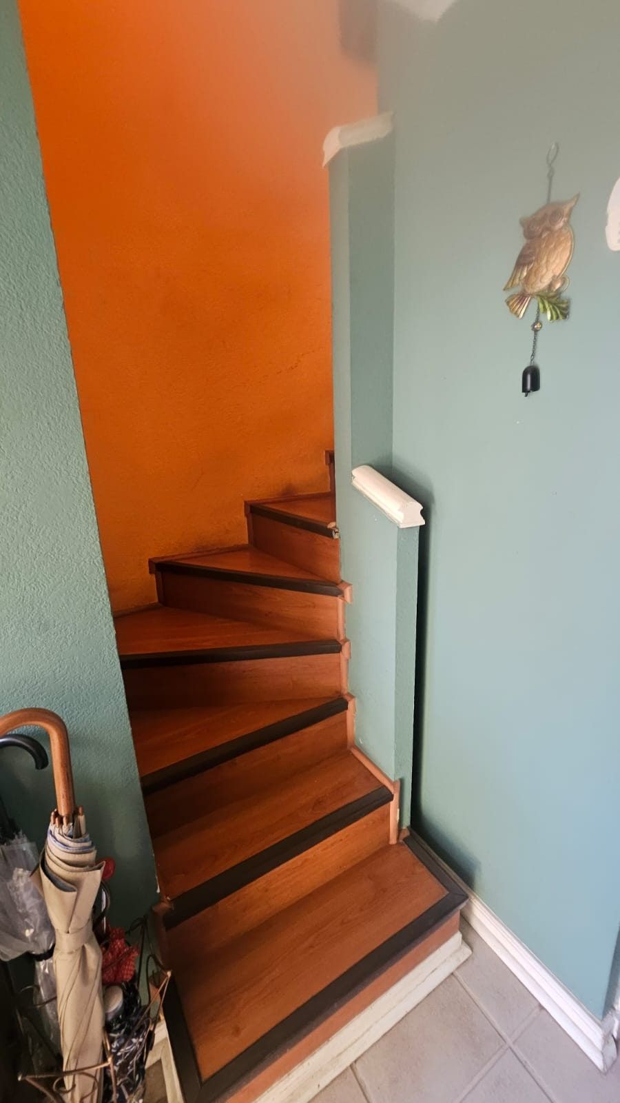 Escalera de madera entre muros naranjo y verde agua, con un adorno de búho colgante en la pared.