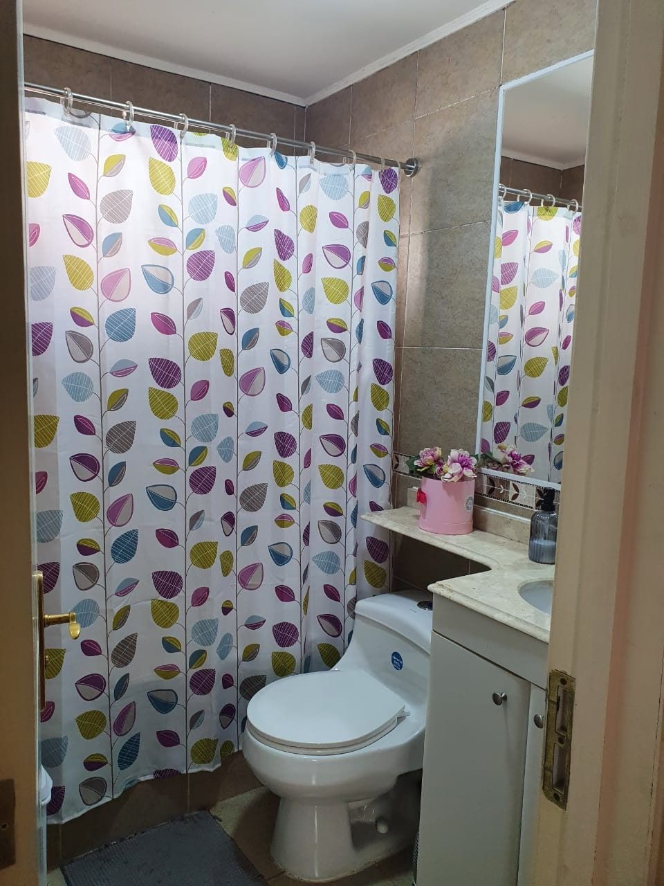 Baño con cortina de ducha estampada, inodoro, mueble de lavamanos con espejo y florero.