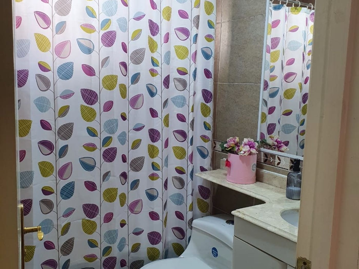 Baño con cortina de ducha estampada, inodoro, mueble de lavamanos con espejo y florero.