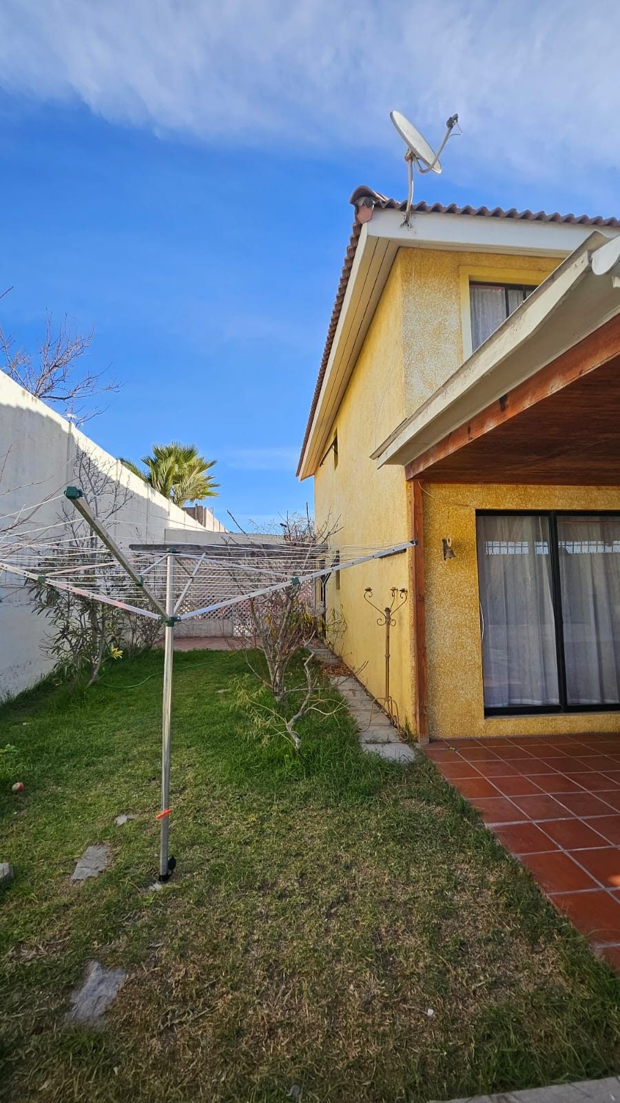 Fachada lateral de la casa amarilla con terraza de baldosas, antena satelital y tendedero de ropa.