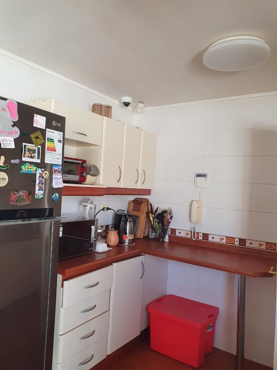 Cocina en L con refrigerador, muebles superiores color crema y barra de madera junto al mesón.