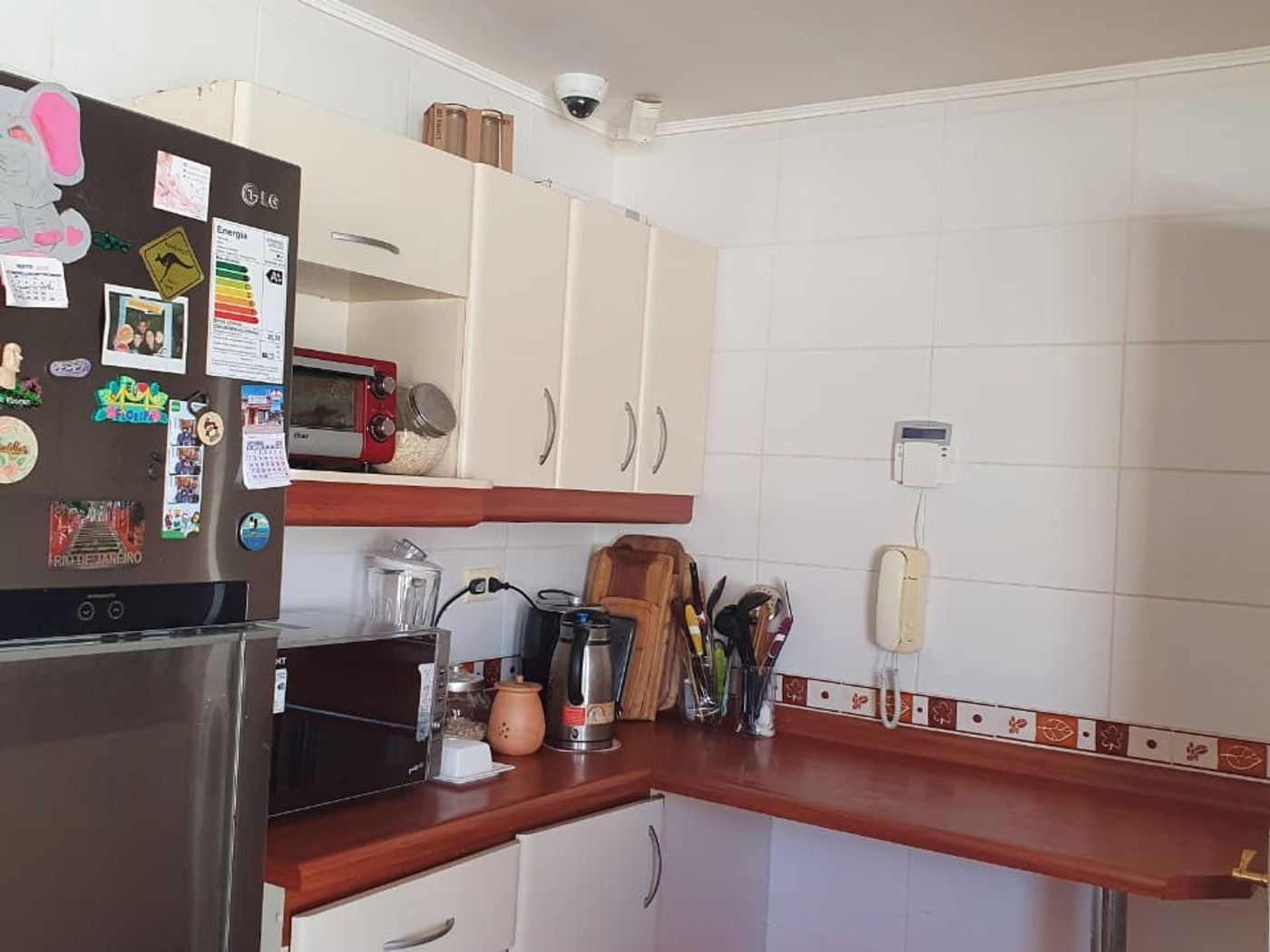 Cocina en L con refrigerador, muebles superiores color crema y barra de madera junto al mesón.