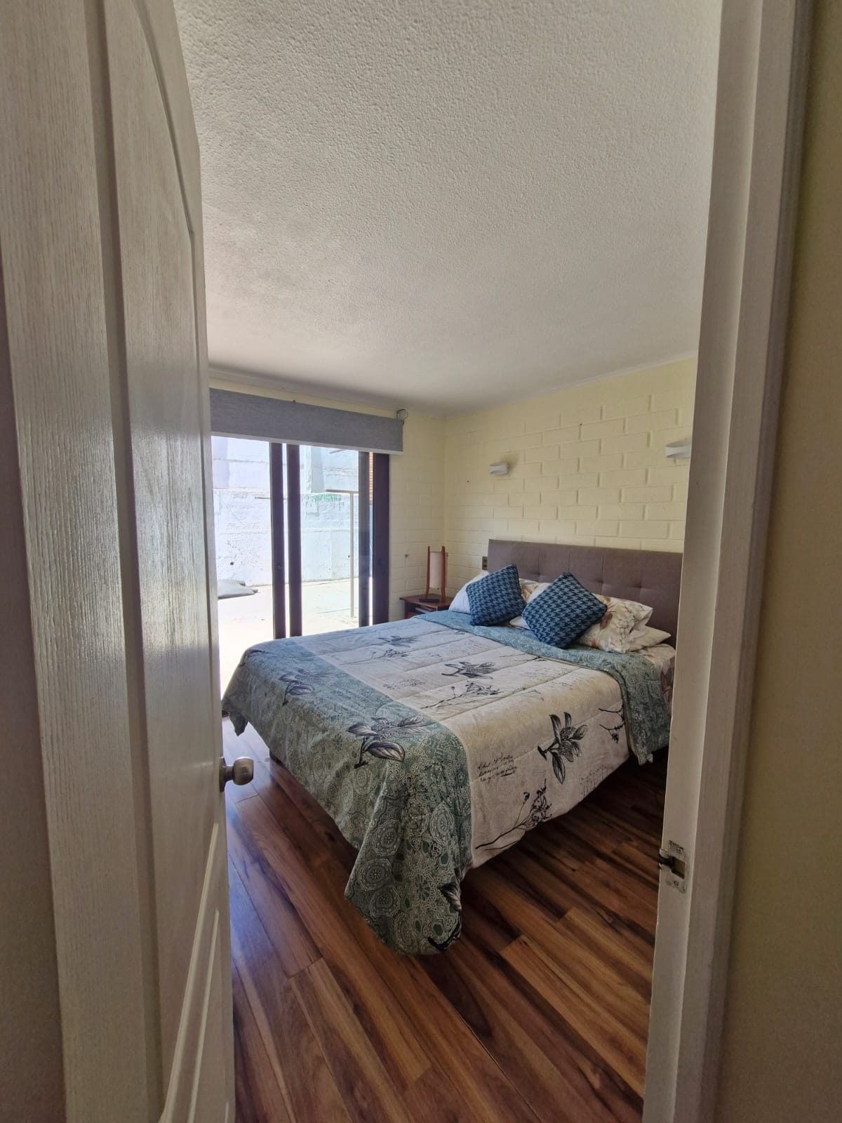 Dormitorio con cama matrimonial, pared de ladrillo pintado y puerta corrediza de vidrio hacia el exterior.
