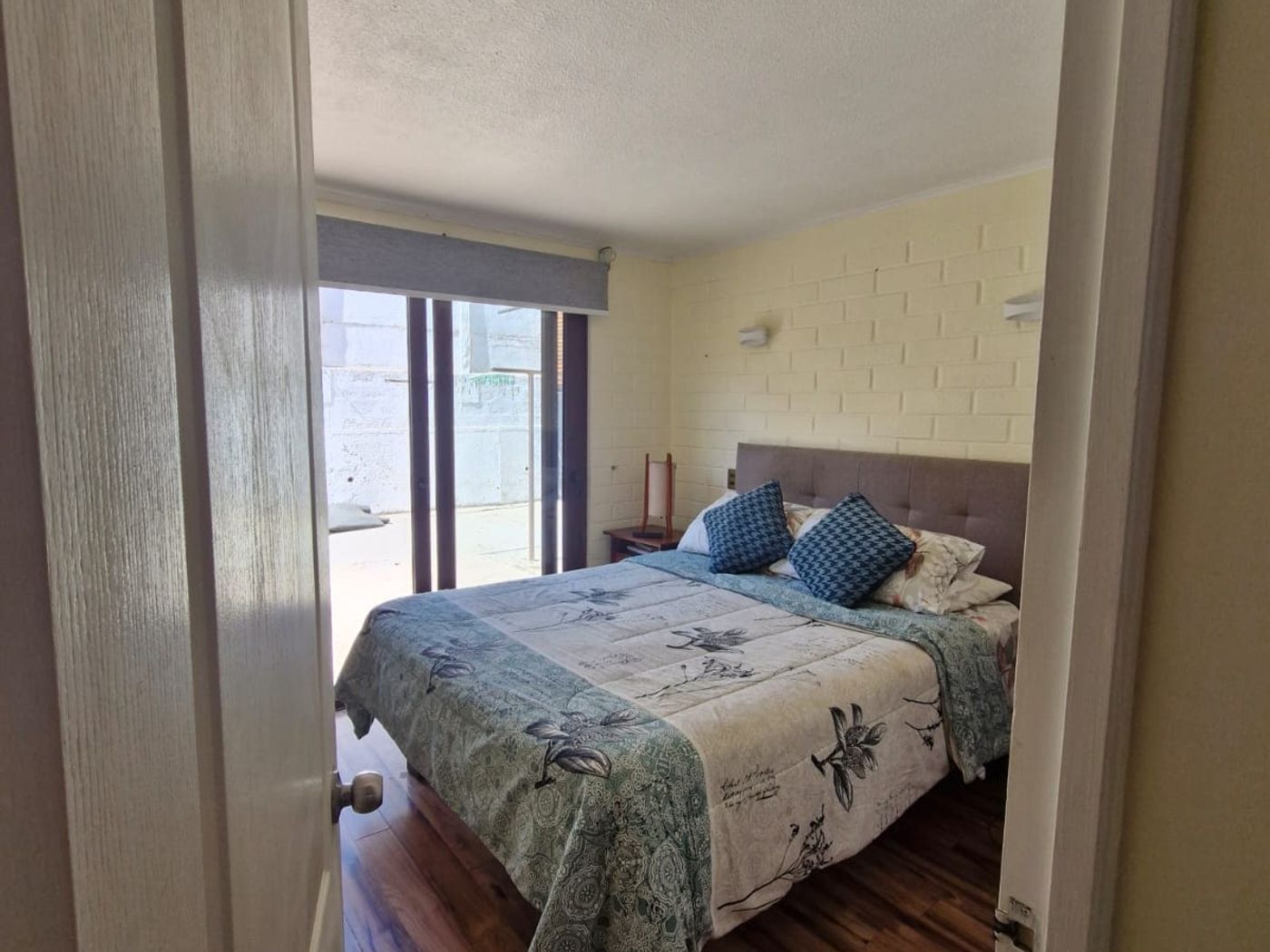 Dormitorio con cama matrimonial, pared de ladrillo pintado y puerta corrediza de vidrio hacia el exterior.