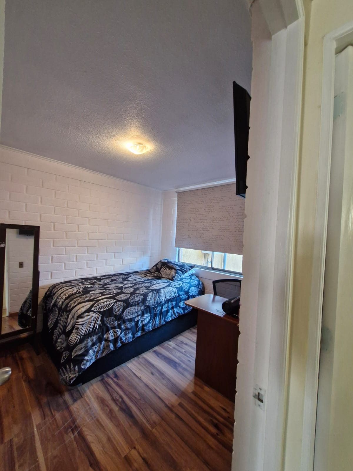 Dormitorio con cama individual, televisor en la pared, escritorio y piso de madera.