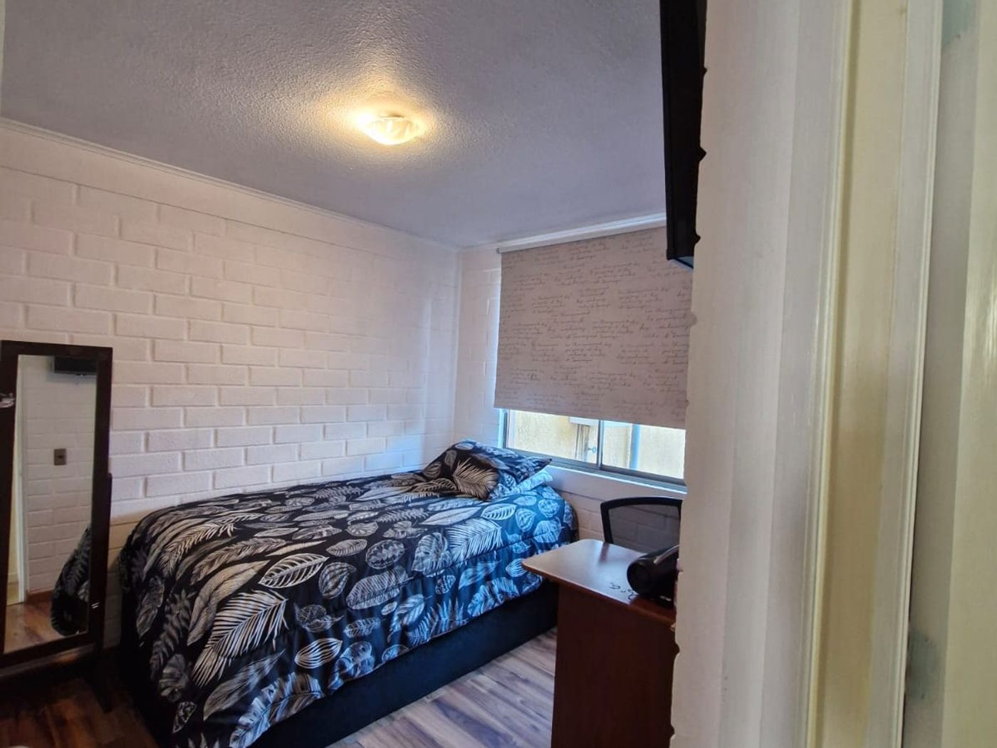 Dormitorio con cama individual, televisor en la pared, escritorio y piso de madera.
