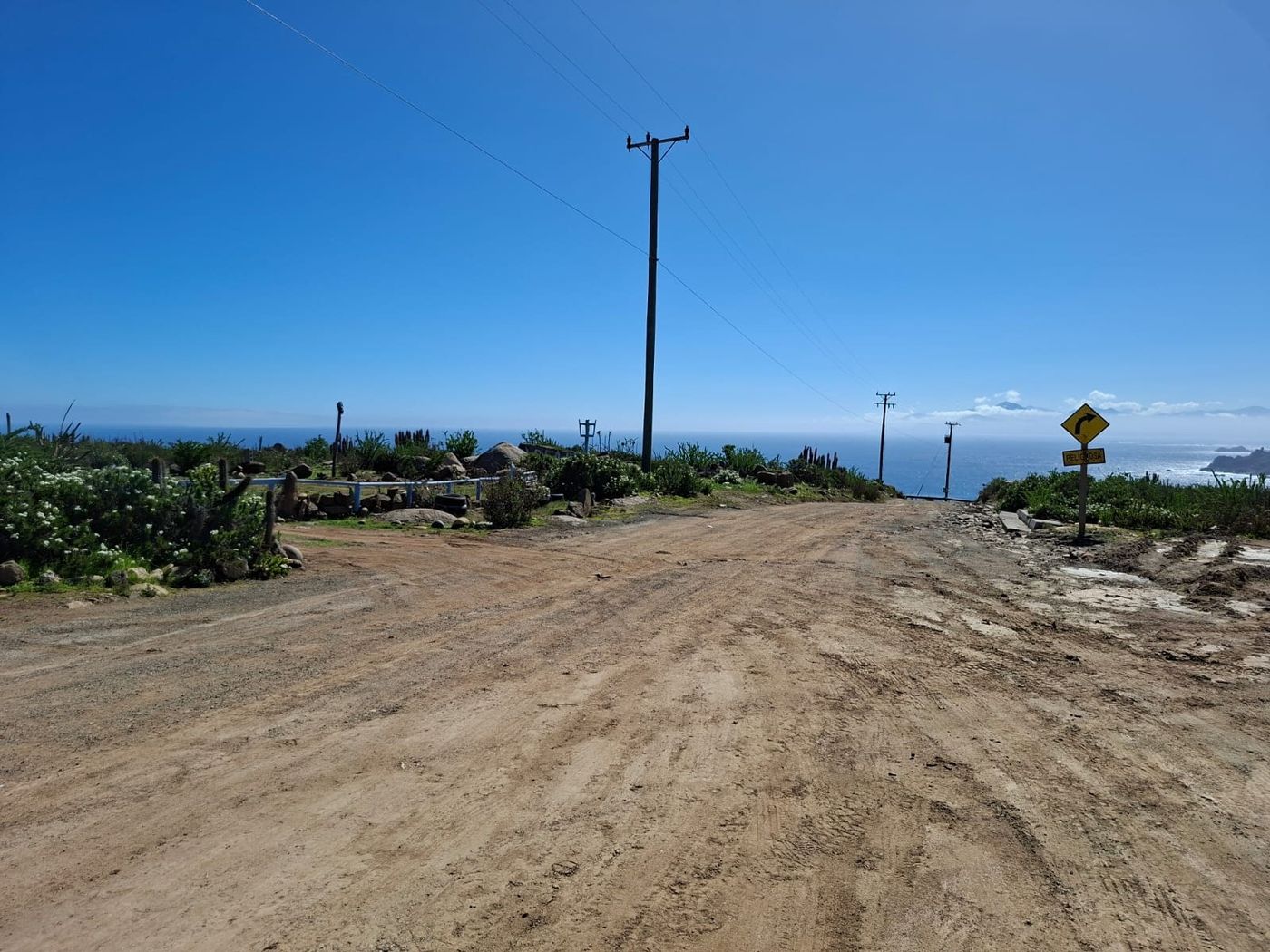 Camino vehicular hacia la costa con señalética de curva y vista al océano Pacífico.