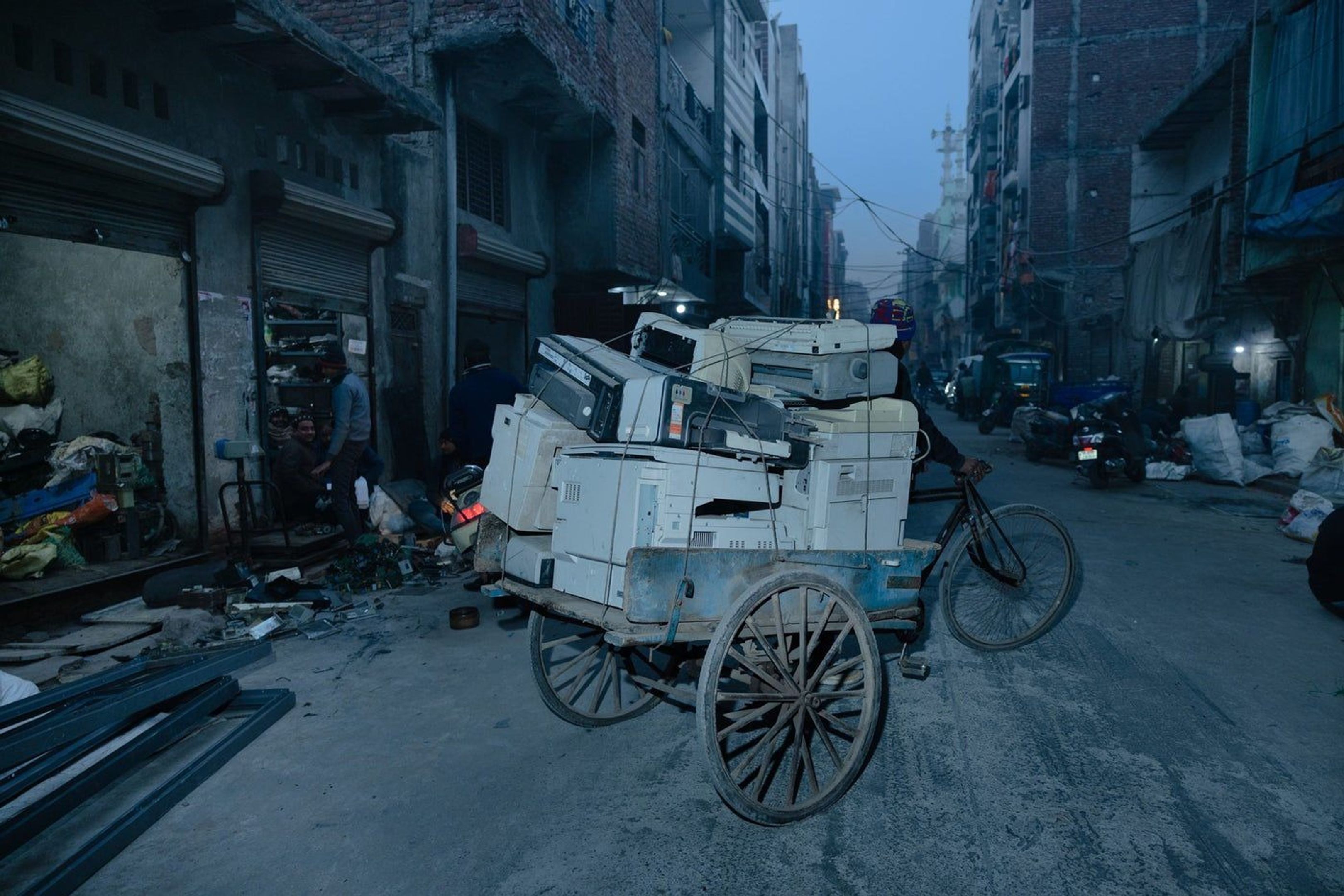 A street scene in a dense urban neighborhood showing a cycle cart or rickshaw heavily loaded with discarded white office electronics, such as old printers and monitors, being transported through a narrow road.