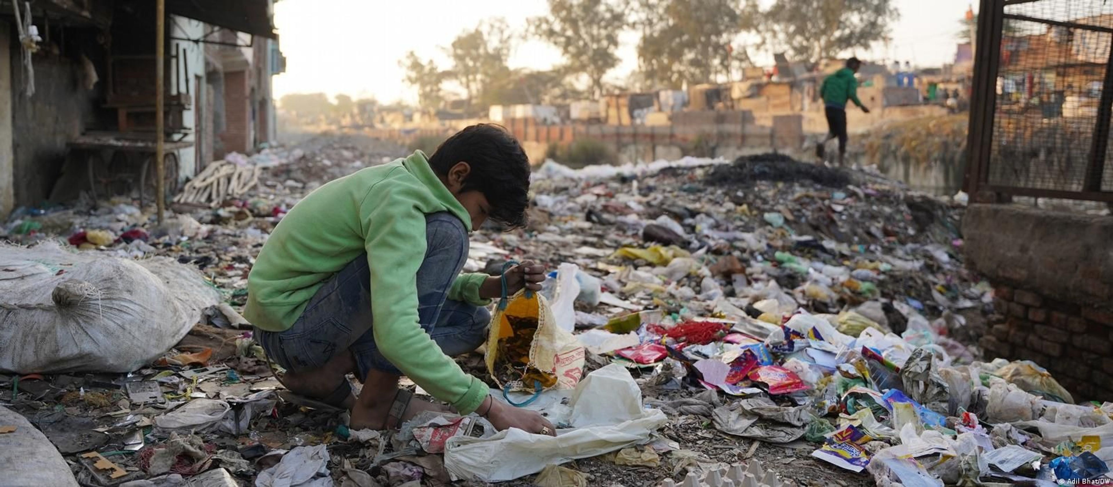 A young boy sits crouched on the ground in a dimly lit workshop, surrounded by large piles of broken laptops, motherboards, and electronic scrap. He is holding a smartphone and using a tool to dismantle it.