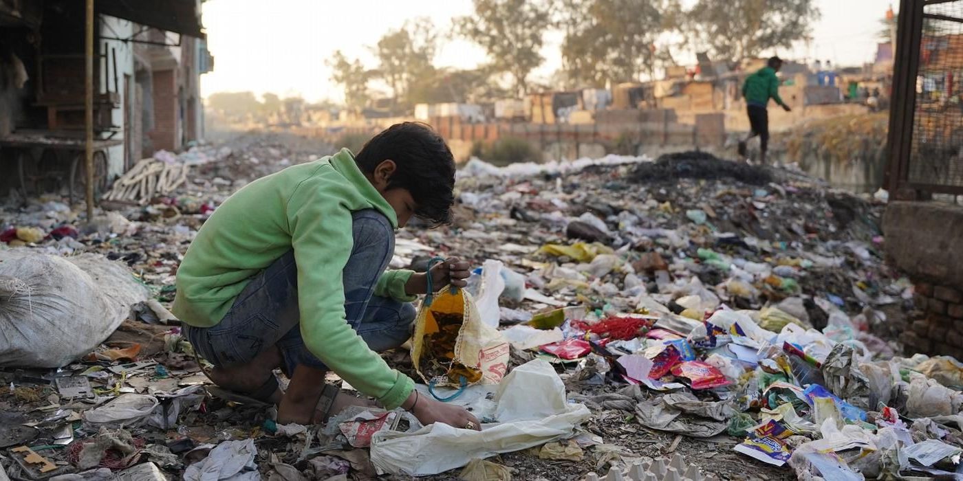 A young boy wearing a green sweatshirt crouches amidst a sprawling landscape of mixed waste and debris, sorting through scraps and placing them into a yellow bag. The background shows a hazy, built-up area typical of informal settlements like Seelampur, with another figure walking along a ridge of refuse in the distance.