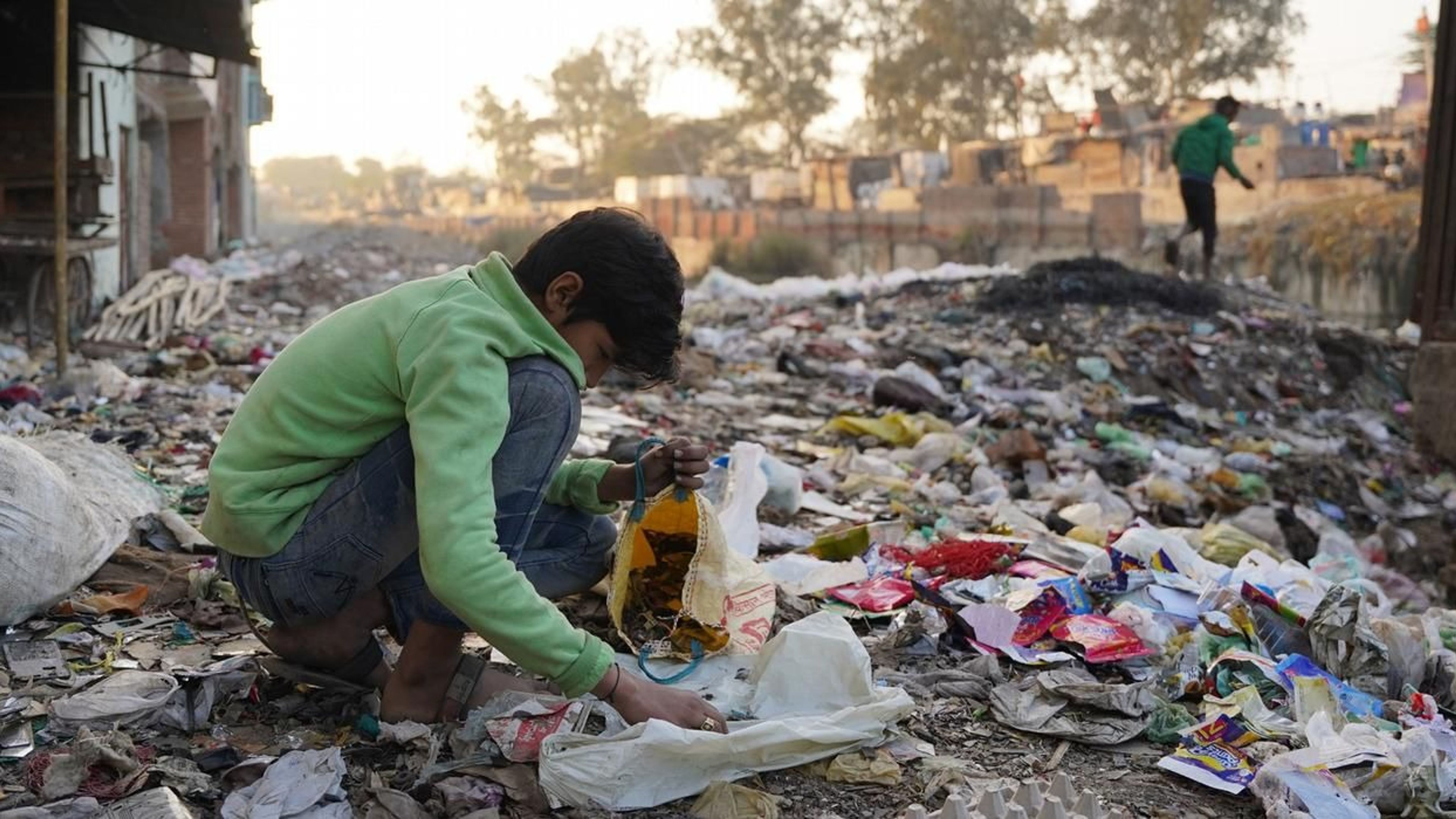 A young boy wearing a green sweatshirt crouches amidst a sprawling landscape of mixed waste and debris, sorting through scraps and placing them into a yellow bag. The background shows a hazy, built-up area typical of informal settlements like Seelampur, with another figure walking along a ridge of refuse in the distance.