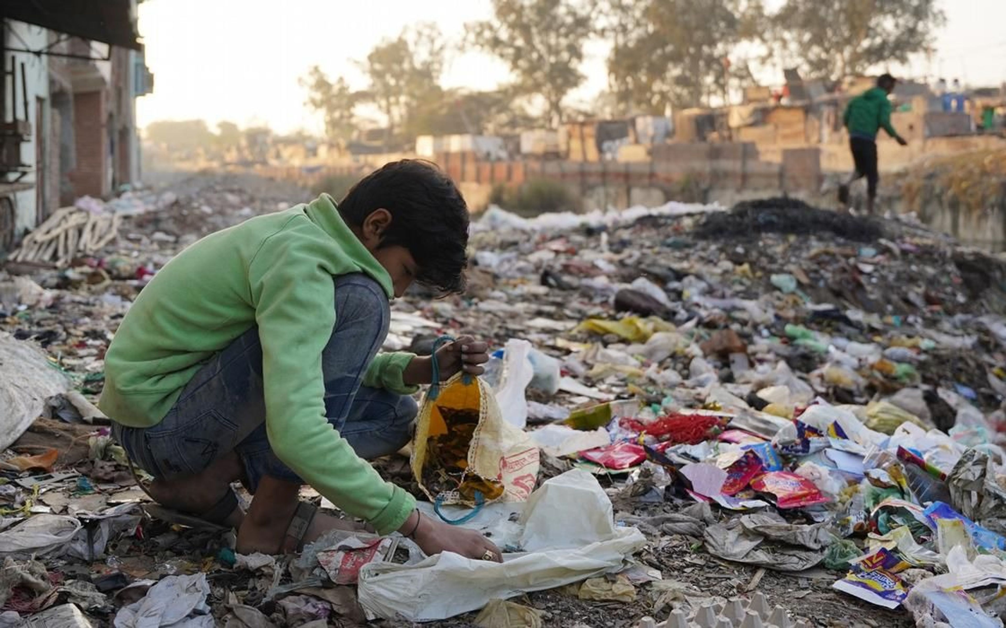A young boy wearing a green sweatshirt crouches amidst a sprawling landscape of mixed waste and debris, sorting through scraps and placing them into a yellow bag. The background shows a hazy, built-up area typical of informal settlements like Seelampur, with another figure walking along a ridge of refuse in the distance.
