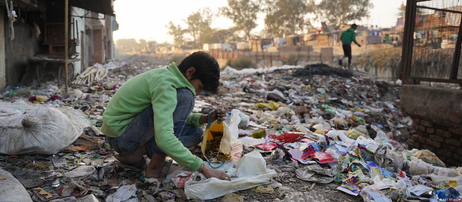 A young boy sits crouched on the ground in a dimly lit workshop, surrounded by large piles of broken laptops, motherboards, and electronic scrap. He is holding a smartphone and using a tool to dismantle it.